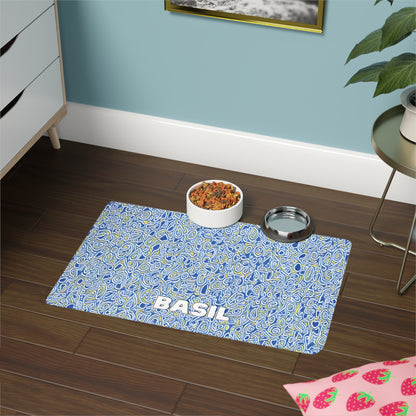 A blue, African-inspired patterned pet placemat with the name BASIL in white letters sits on a dark wood floor. On the mat are a bowl of kibble and a metal water bowl. Nearby are a green plant, white dresser, and strawberry-patterned mat.