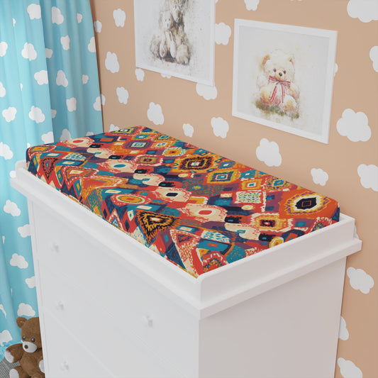 A white changing table features a jersey knit changing pad cover in vibrant red, blue, and orange tones. Cloud wallpaper, framed bear illustrations, and a blue curtain with white clouds brighten the space. A brown teddy bear sits by the dresser.