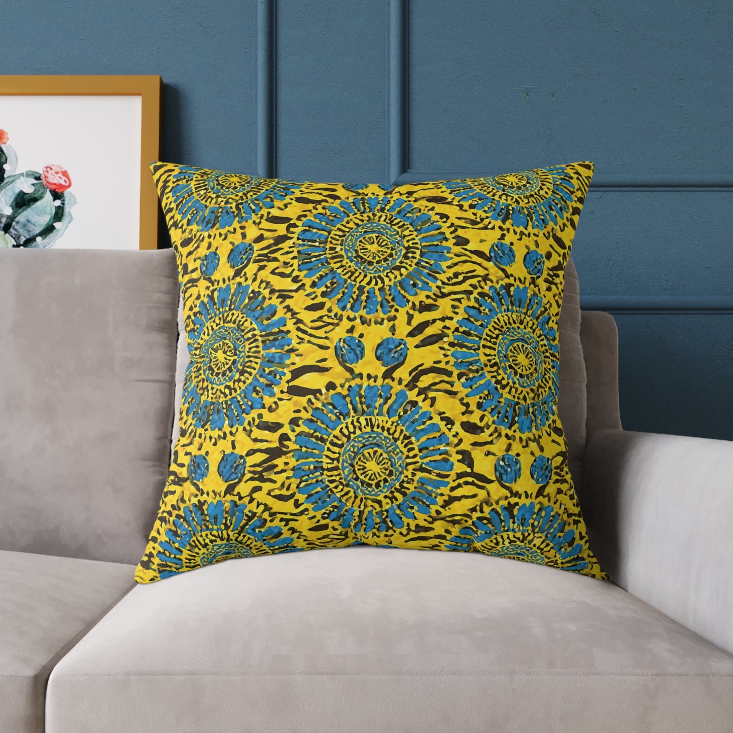 A vibrant throw pillow with a bold yellow background and blue mandala-like floral patterns sits on a light gray sofa against a dark blue wall. A framed artwork with green and red cacti is partially visible in the background to the left.