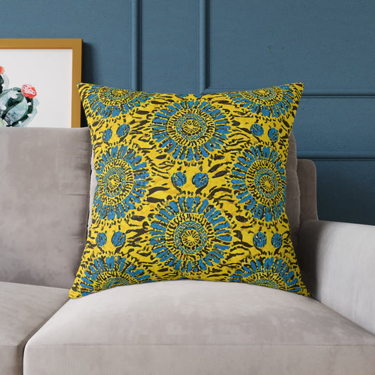 A vibrant throw pillow with a bold yellow background and blue mandala-like floral patterns sits on a light gray sofa against a dark blue wall. A framed artwork with green and red cacti is partially visible in the background to the left.
