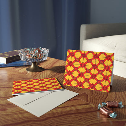 A greeting card and envelope with a bold red and yellow, African-inspired geometric pattern sit on a wooden table. Nearby are wrapped chocolates, an open book, and a glass candy dish. A gray couch and blue curtain are in the background.