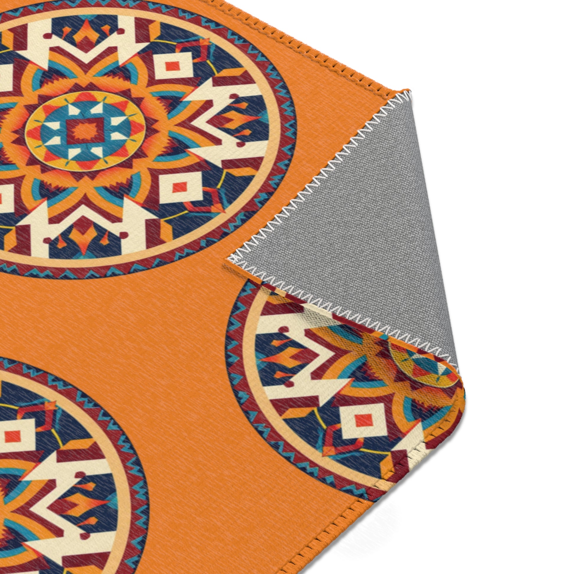 A rectangular fabric with an orange background features African-inspired circular, geometric patterns in blue, red, yellow, and cream. The fabric corner is flipped to show a gray, textured underside and zigzag stitching along the edge on a white background.