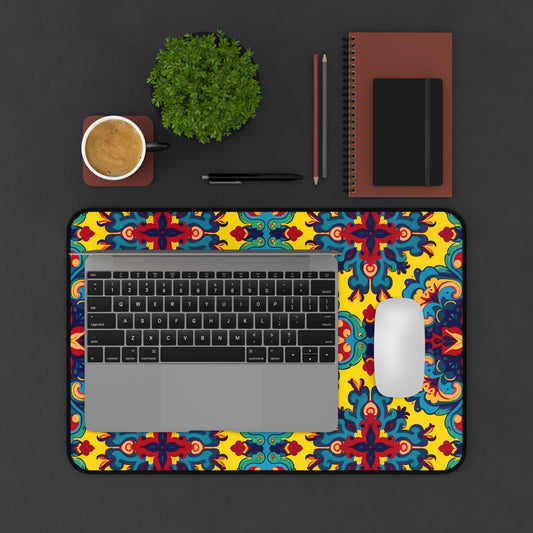 A laptop and white mouse sit on a colorful neoprene desk mat with red, blue, and yellow designs. Nearby are a brown notebook, writing tools, a small green potted plant, and coffee on a brown saucer atop the dark surface.