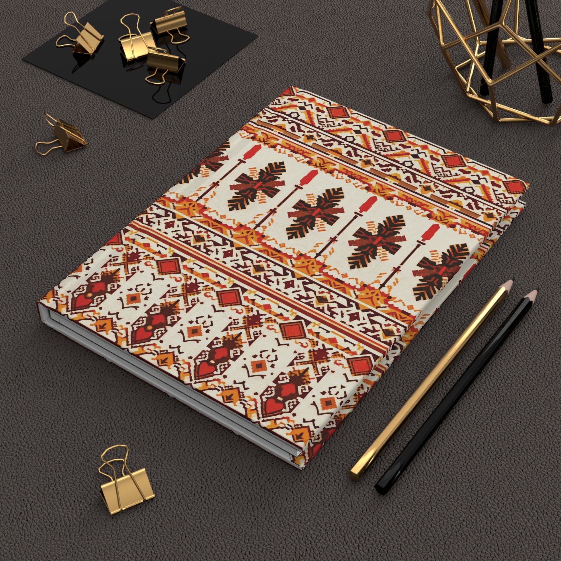 A tribal pattern notebook with red, orange, black, and cream geometric designs lies on a textured brown surface. Nearby are a gold and a black pencil, gold binder clips, and a decorative geometric metal sculpture.