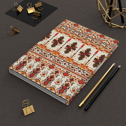 A tribal pattern notebook with red, orange, black, and cream geometric designs lies on a textured brown surface. Nearby are a gold and a black pencil, gold binder clips, and a decorative geometric metal sculpture.