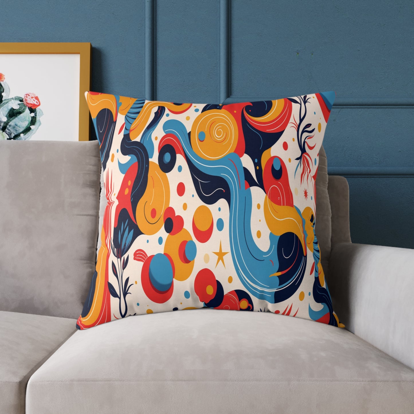 A colorful accent pillow with an abstract design featuring swirling lines in blue, yellow, orange, and red rests on a light gray sofa. Perfect for vibrant home decor, it pops against the blue wall and a framed painting with green and red on the left.