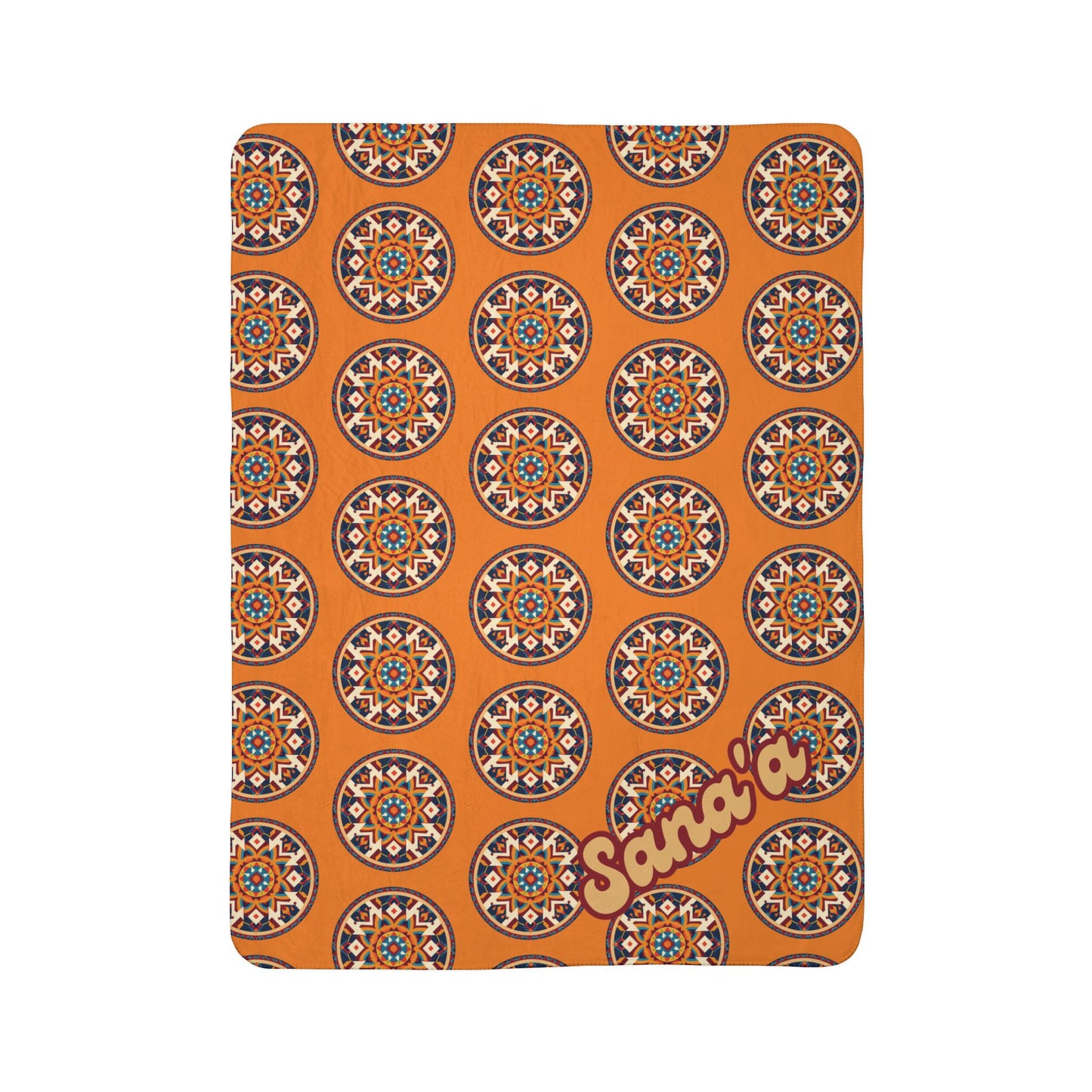 A rectangular orange blanket with a repeating African-inspired circular geometric pattern in blue, white, and red. In the lower right corner, Sana’a is written in large, light yellow and dark red outlined script. The background is plain white.