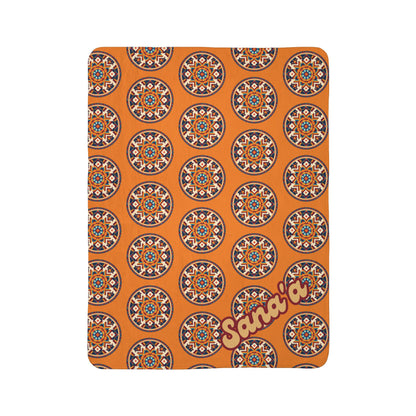 A rectangular orange blanket with a repeating African-inspired circular geometric pattern in blue, white, and red. In the lower right corner, Sana’a is written in large, light yellow and dark red outlined script. The background is plain white.