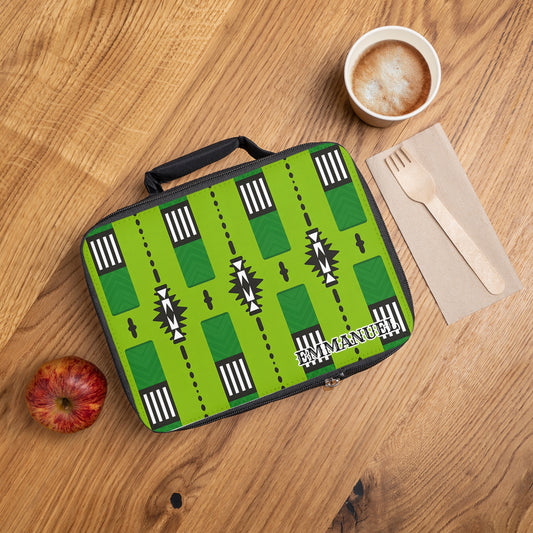 A green African-inspired lunch bag personalized with the name Emmanuel sits on a wooden table beside a red apple, a cup of cappuccino with foam art, and a napkin holding a wooden fork and knife.