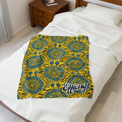 A vibrant yellow and blue African-inspired blanket with a bold, circular floral design and “Effiong” in white script lies neatly over a white blanket on a made bed. Beside the bed, a wooden nightstand holds two books.