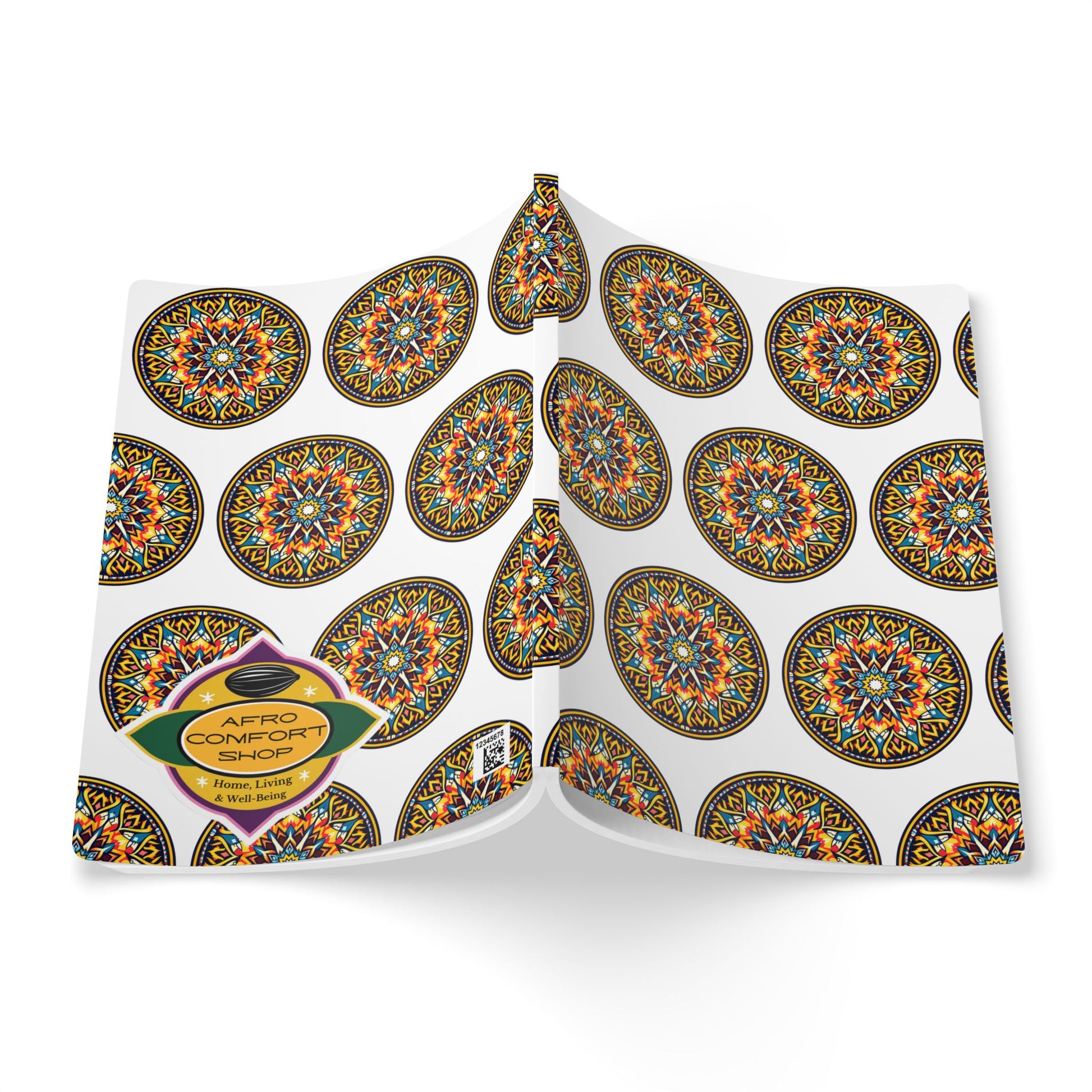 A hardcover African-inspired journal lies open, its white cover adorned with colorful, circular geometric patterns. An oval label at the bottom left reads “Afro Comfort Shop” and “Handmade Items” in orange and yellow on a dark background.