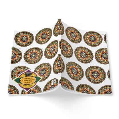 A hardcover African-inspired journal lies open, its white cover adorned with colorful, circular geometric patterns. An oval label at the bottom left reads “Afro Comfort Shop” and “Handmade Items” in orange and yellow on a dark background.