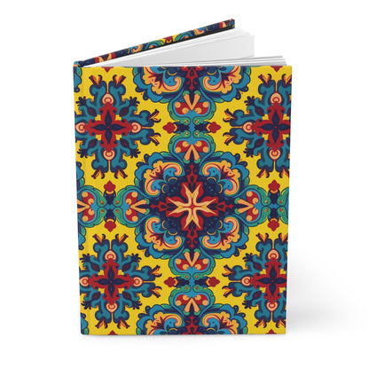 An African-inspired hardcover journal features an ornate, symmetrical floral and geometric pattern in red, blue, teal, and orange on a bright yellow background. The notebook stands upright and slightly open, revealing crisp white pages inside.