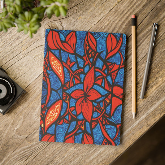 An African-inspired journal with a bold abstract floral cover in red, blue, and orange rests on a wooden table. Beside it are a pencil, a silver pen, a camera, and the edge of a potted plant visible in the corner.
