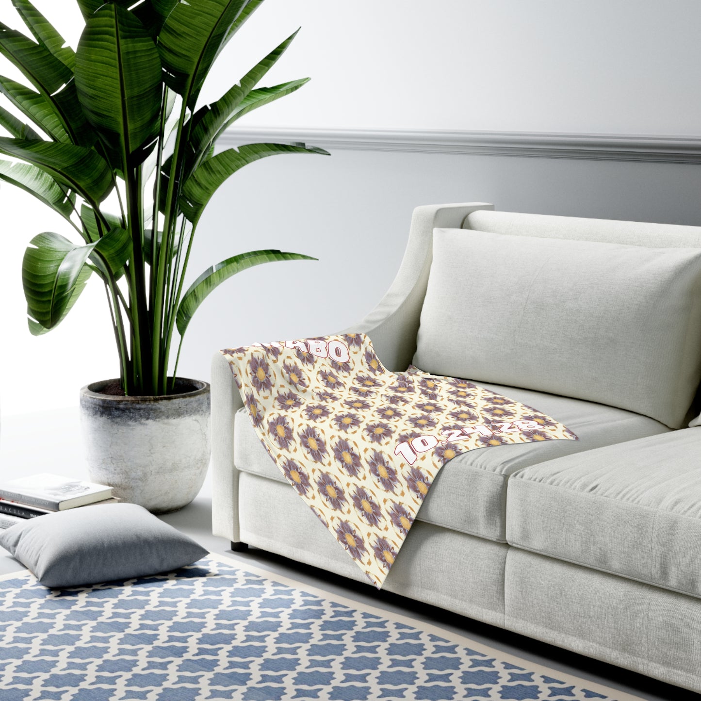 A modern living room with a light gray sofa draped with a patterned blanket, near a large green potted plant. African-inspired decor accents complement the blue and white geometric rug, adding character to the room’s minimal, bright aesthetic.