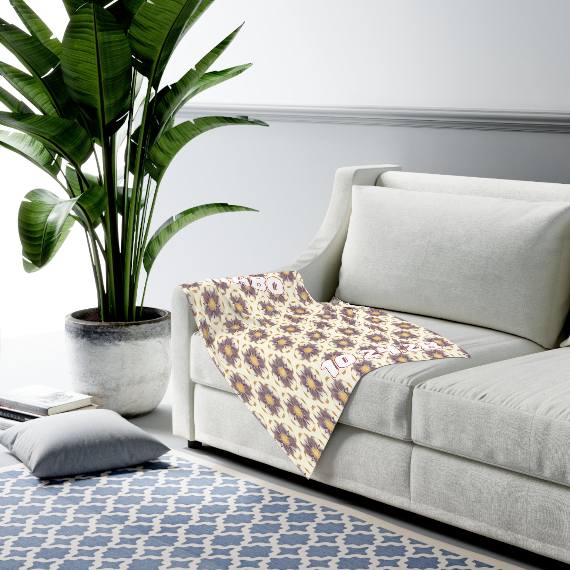 A modern living room with a light gray sofa draped with a patterned blanket, near a large green potted plant. African-inspired decor accents complement the blue and white geometric rug, adding character to the room’s minimal, bright aesthetic.