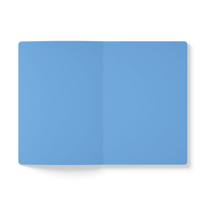 A top-down view of an open creative journal with blank, light blue pages. The journal lies flat on a white background, featuring a visible vertical crease in the center where the pages meet. No text, lines, or images appear on the pages.