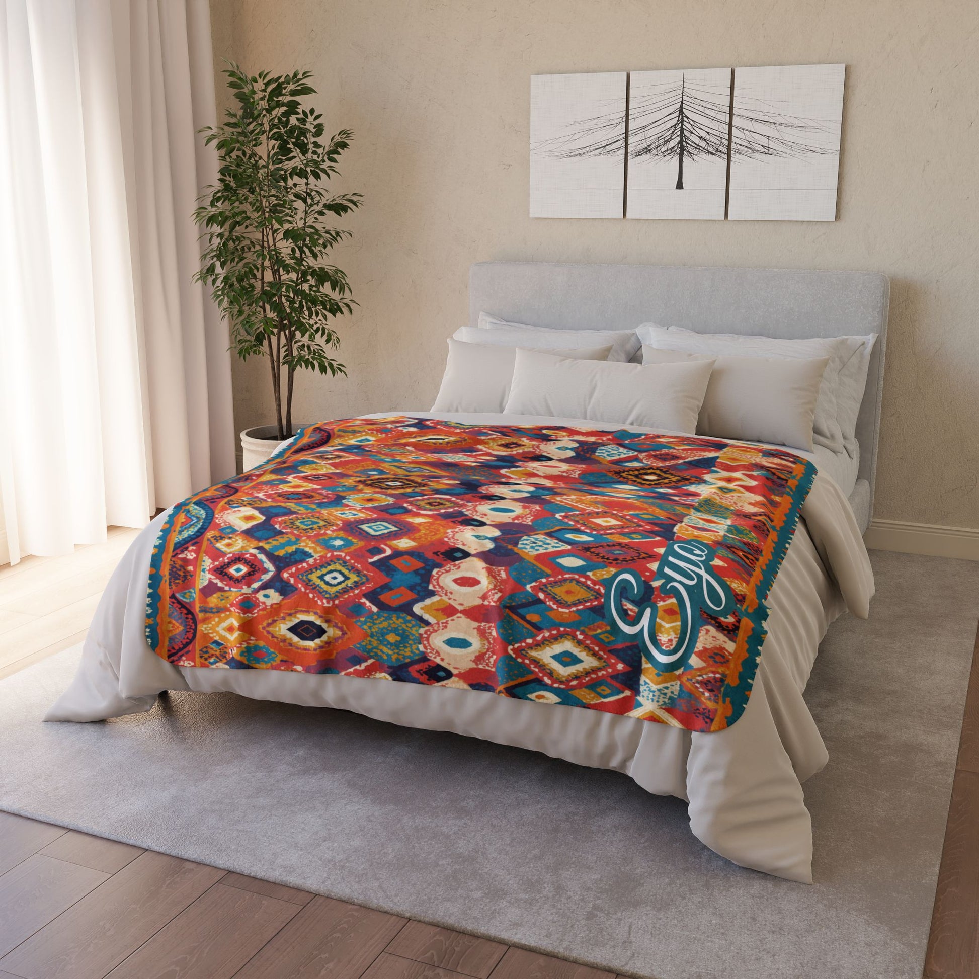 A neatly made bed with white bedding and an African-inspired geometric-patterned blanket in vibrant reds, blues, and yellows. The room features a gray headboard, white pillows, a green potted plant, and minimalist wall art above the bed.