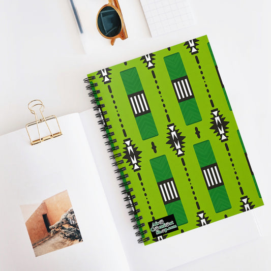 A green African-inspired spiral notebook with a bold black and white geometric pattern lies on a white desk. Nearby are an open book with a photo, gold binder clip, round sunglasses, and a small white object, creating a bright, creative workspace.