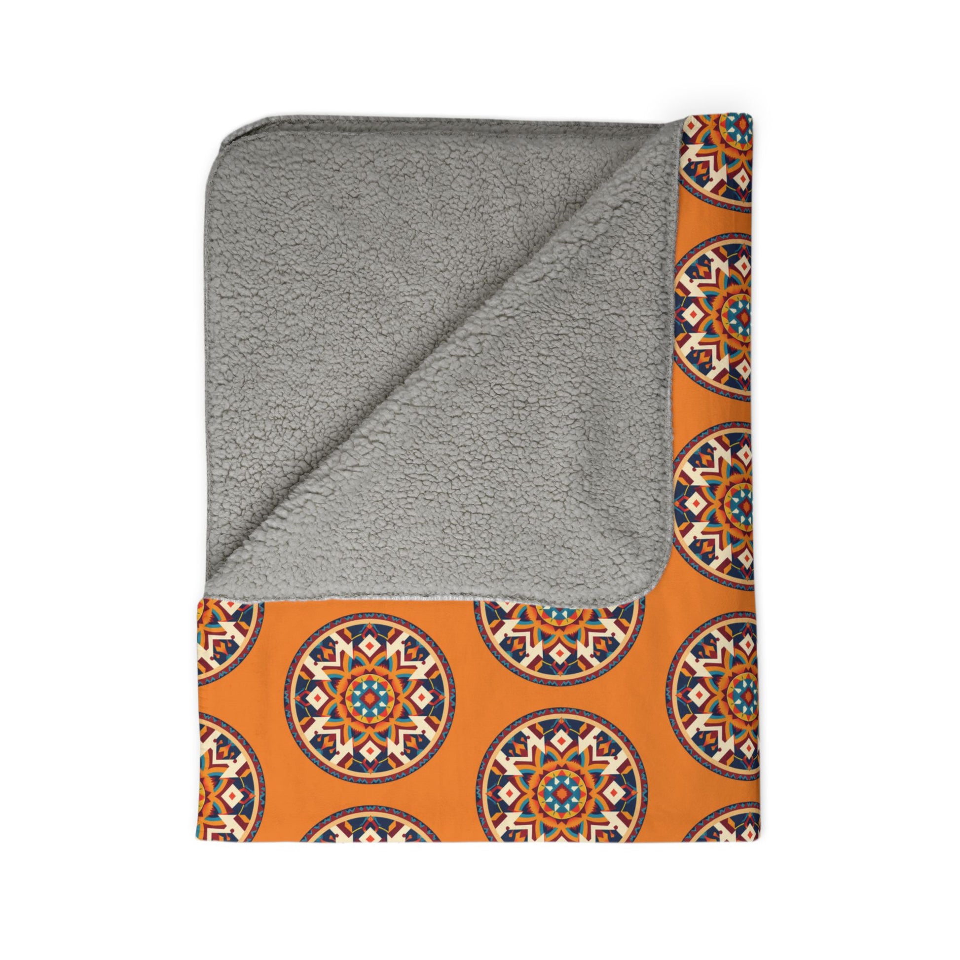 A folded blanket with a soft, light gray fleece underside and an orange fabric top featuring a repeating African-inspired circular geometric mandala pattern in blue, black, and white. One corner is flipped to show the fleece lining.