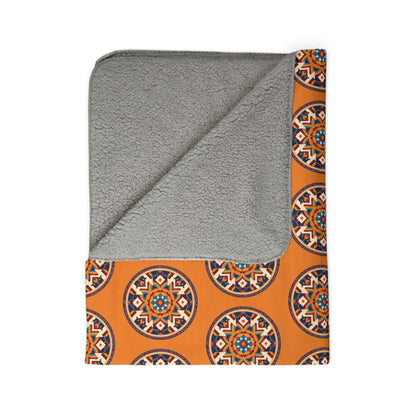 A folded blanket with a soft, light gray fleece underside and an orange fabric top featuring a repeating African-inspired circular geometric mandala pattern in blue, black, and white. One corner is flipped to show the fleece lining.