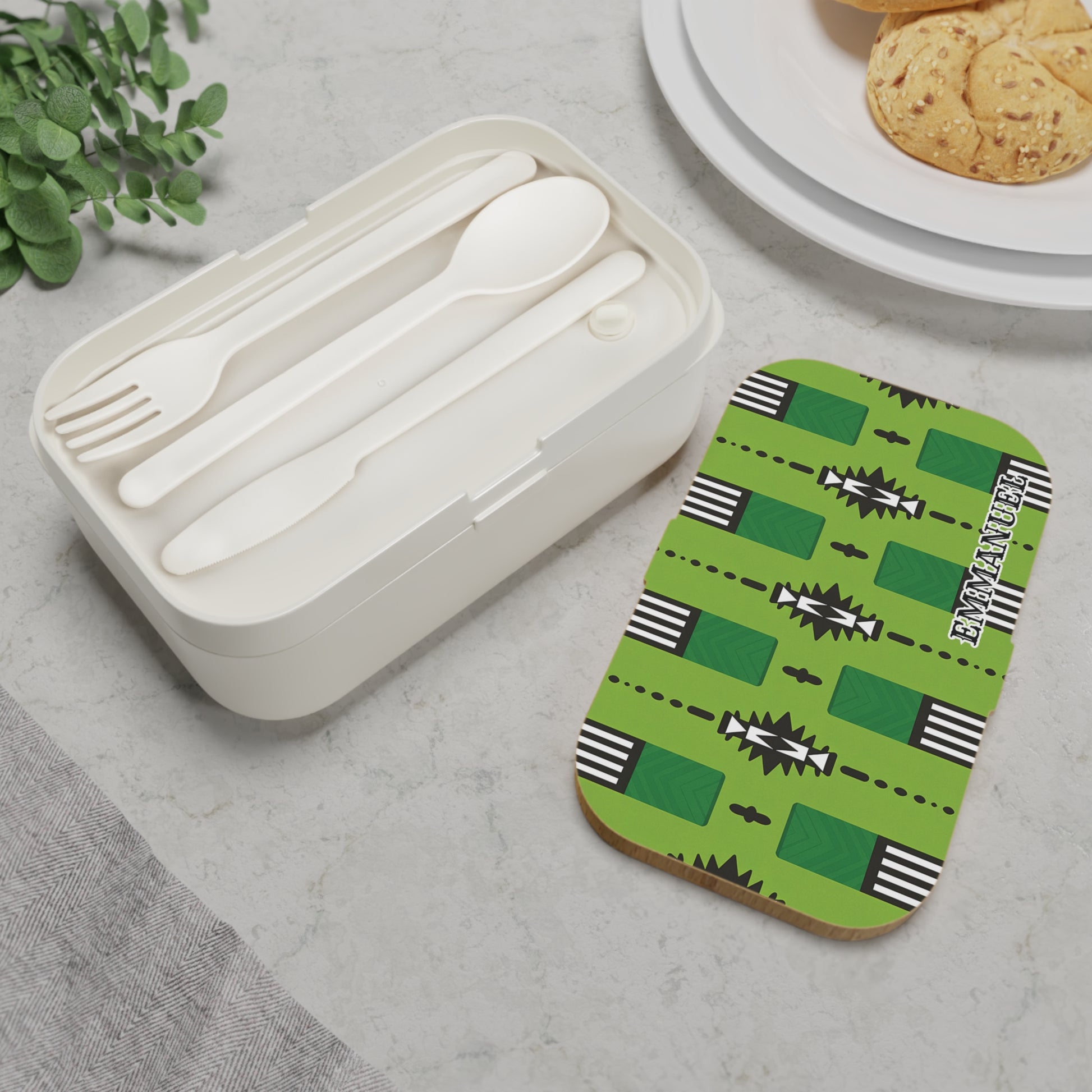A white personalized bento lunch box with a fork, spoon, and knife sits on marble. Its lid features a bold green geometric pattern and “ETHNIC DESIGN.” Nearby are cookies on a white plate and a small plant.