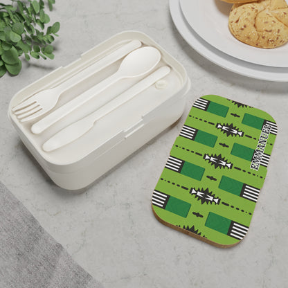 A white personalized bento lunch box with a fork, spoon, and knife sits on marble. Its lid features a bold green geometric pattern and “ETHNIC DESIGN.” Nearby are cookies on a white plate and a small plant.