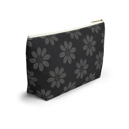 A black zippered cosmetic travel bag with a gold zipper is shown. The pouch features a repeating pattern of large, hand-drawn light gray daisies, inspired by African-inspired patterns, on a solid black background and rests on a white surface.