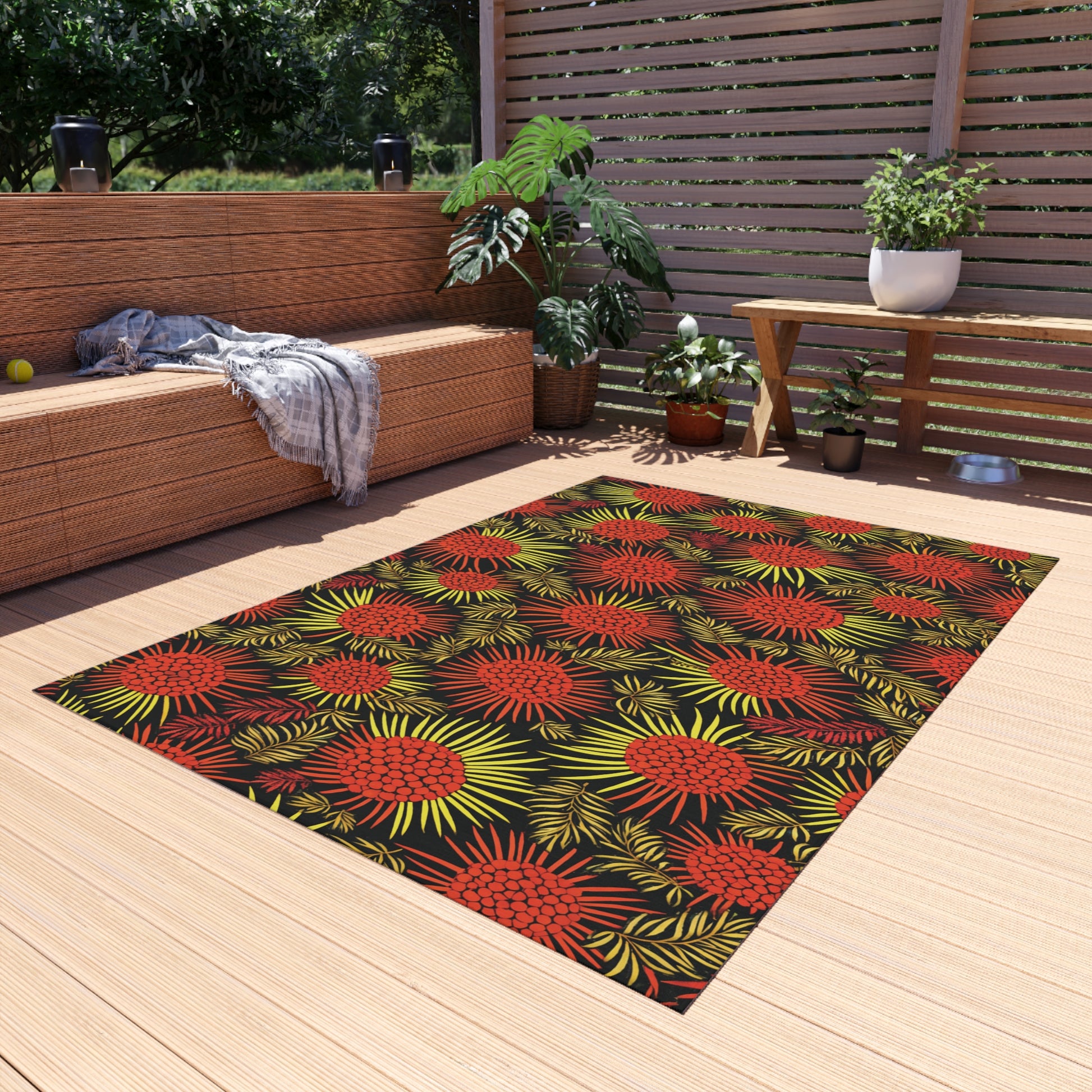 A modern patio with wooden decking features an African-inspired, colorful rug with red and yellow floral patterns. There’s a wicker sofa with cushions and a gray throw, potted plants, and a wooden table. Sunlight filters through a slatted privacy screen.