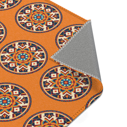 An orange fabric features repeating African-inspired, mandala-like patterns in blue, red, white, and yellow. One corner is folded over, revealing a gray underside with white zigzag stitching along the edge.