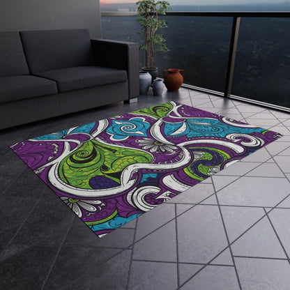 A modern balcony with a dark gray tile floor features a weather-resistant rug in swirling abstract patterns of purple, green, blue, and white. A gray sofa sits on the left, while a potted plant rests near the glass railing overlooking the cityscape.