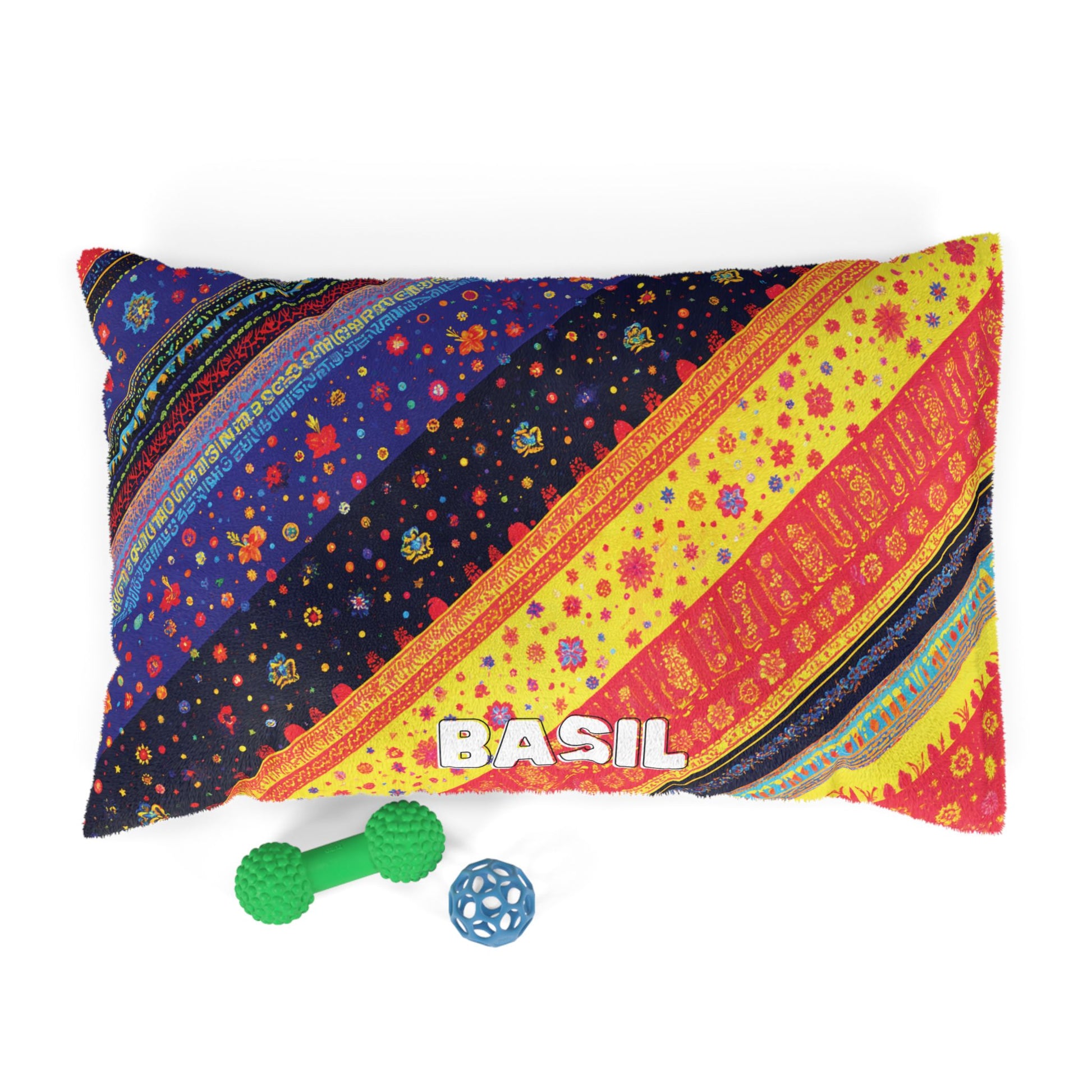 A rectangular pillow with colorful, African-inspired diagonal stripes and floral patterns, featuring the word “BASIL” in white text. In front, there’s a green textured dumbbell-shaped dog toy and a blue ball-shaped toy with geometric openings.