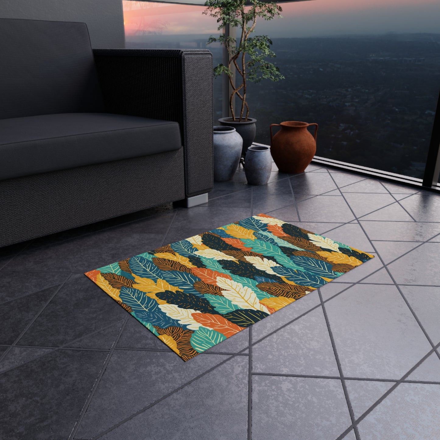 A colorful African-inspired leaf-patterned rug lies on a tiled balcony floor. Next to it are potted plants and clay pots. Part of a dark gray sofa is visible. The balcony overlooks a city and hills at sunset, framed by an orange and blue sky.