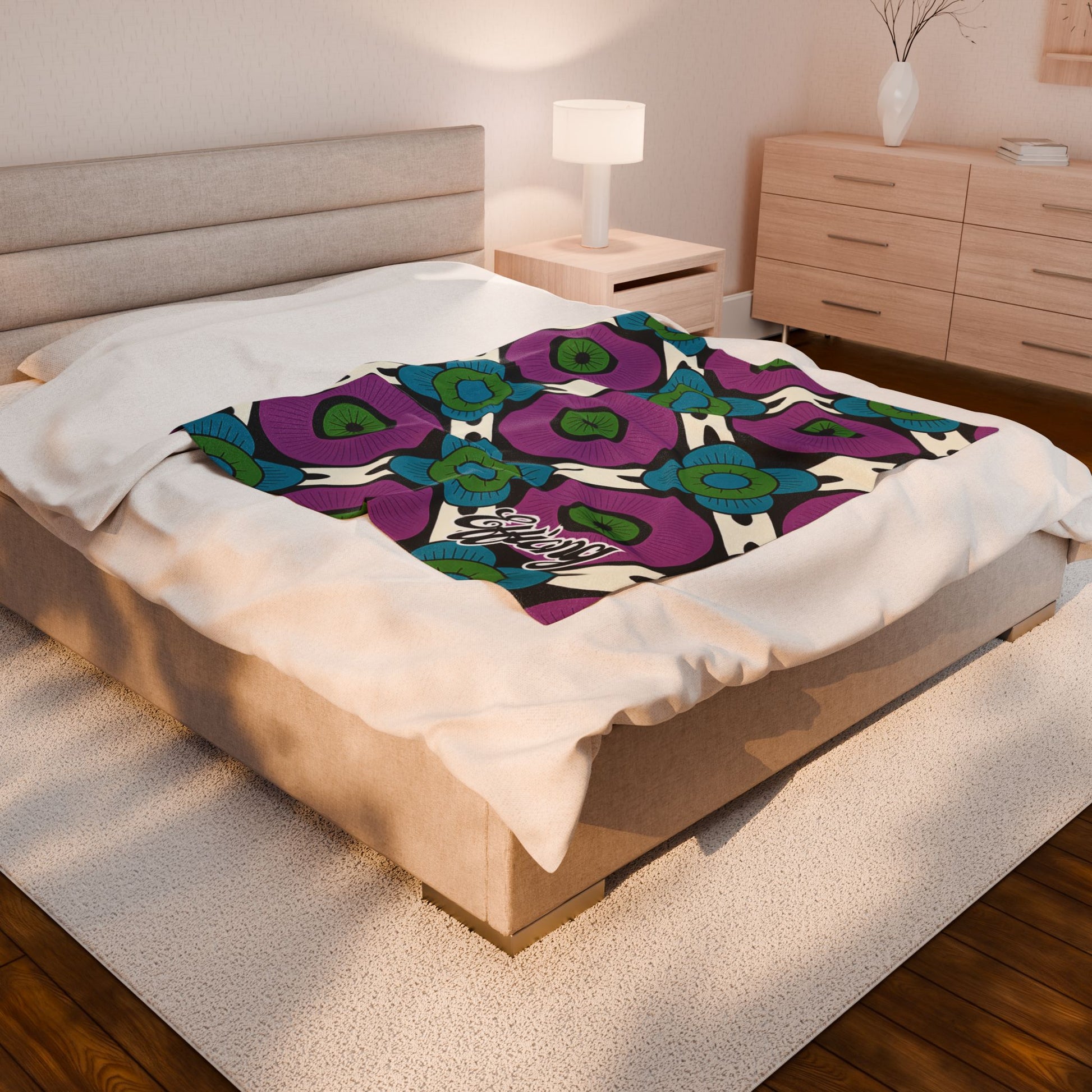 A beige bed with white linens features an African-inspired blanket with a bold floral pattern in purple, blue, and green. The room has a light wood nightstand, lamp, dresser, vase with twigs, and a textured cream rug on a wood floor.