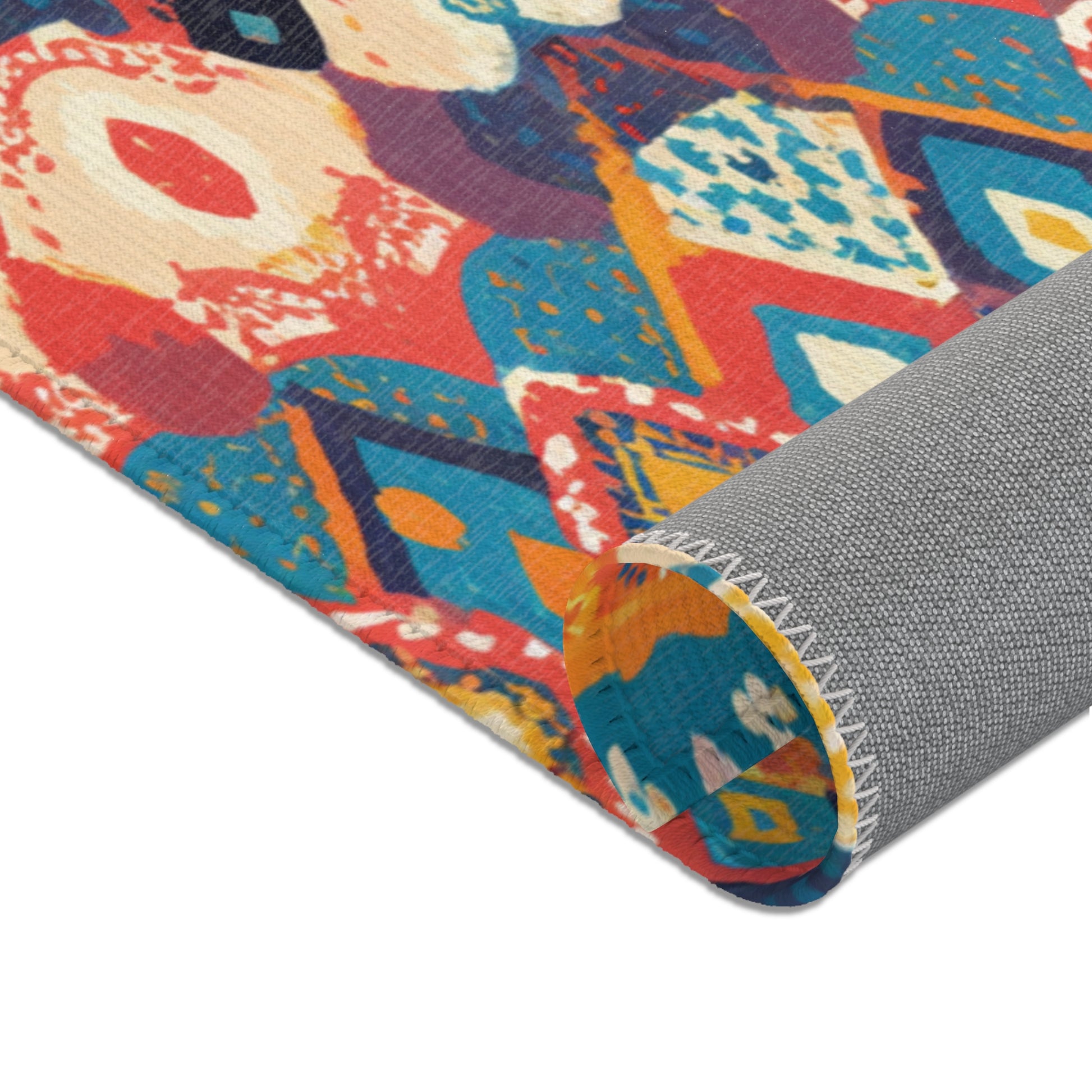 A close-up of a colorful rug being rolled up, revealing an abstract geometric pattern in red, blue, yellow, orange, and white. Made from durable polyester rugs material, the edge is stitched and the plain gray underside features visible border stitching.
