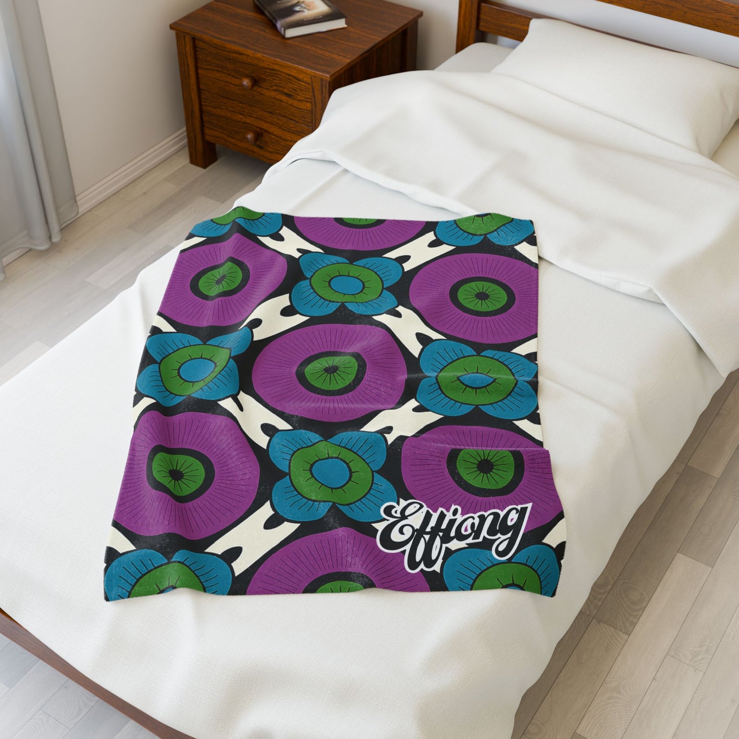 A white bed is covered with an African-inspired personalized blanket, featuring a bold floral pattern of large purple and blue flowers with green centers. The word Effiong appears in white script. The bed is beside a wooden nightstand and a window.