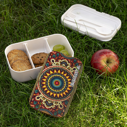 A BPA-free lunchbox with a colorful, intricate mandala design sits open on green grass. Inside, cookies and a green grape rest in compartments. Nearby are a red apple and the white container lid holding a fork and spoon.