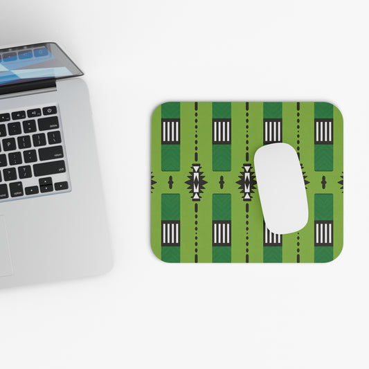 A silver laptop sits open on a white surface next to a vibrant desk accessory: an African-inspired mouse pad with a non-slip rubber base, featuring black geometric and white striped patterns. A white mouse rests on the pad. The composition is minimal and modern.