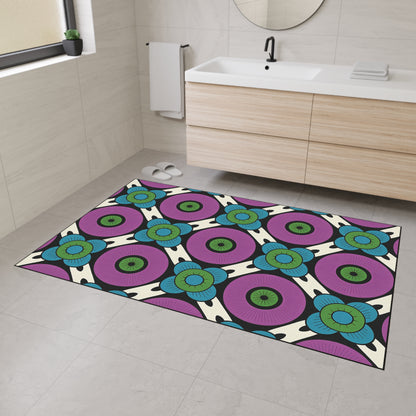 A modern bathroom with light-colored tiled walls and floor, a wooden vanity, and a round mirror features a colorful, African-inspired custom floor mat with a bold geometric pattern in purple, blue, green, and black and non-slip rubber backing.