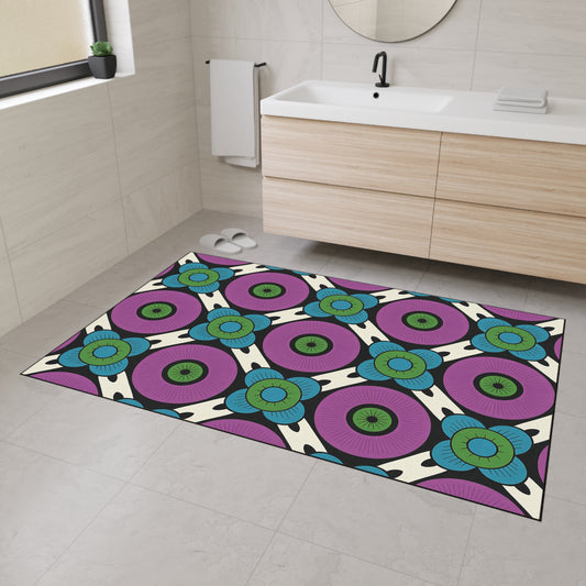 A modern bathroom with light-colored tiled walls and floor, a wooden vanity, and a round mirror features a colorful, African-inspired custom floor mat with a bold geometric pattern in purple, blue, green, and black and non-slip rubber backing.