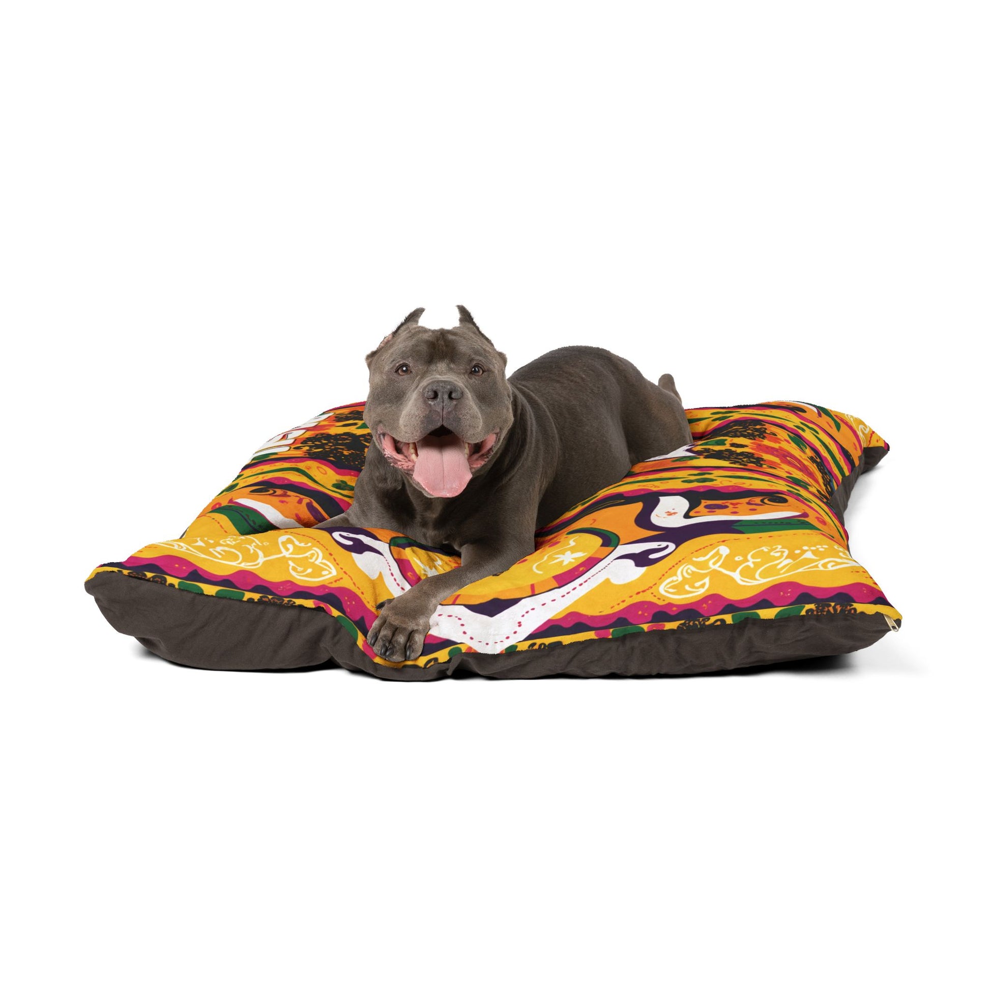 A happy gray pit bull with cropped ears lies on a colorful, African-inspired dog bed. The dog has its mouth open, tongue out, and looks directly at the camera. The bed features bright orange, yellow, purple, and black abstract designs.