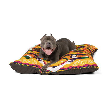 A happy gray pit bull with cropped ears lies on a colorful, African-inspired dog bed. The dog has its mouth open, tongue out, and looks directly at the camera. The bed features bright orange, yellow, purple, and black abstract designs.