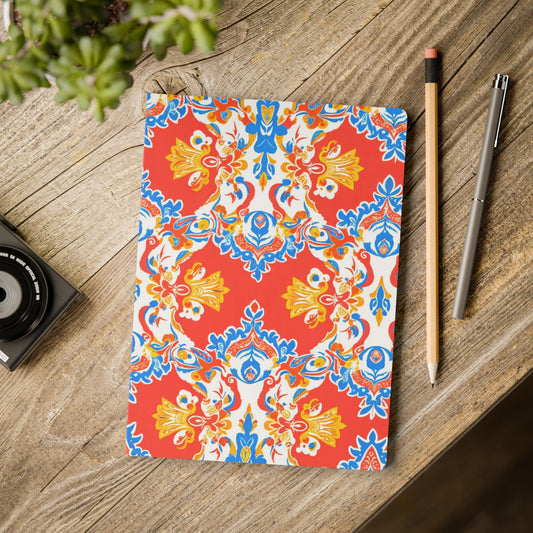 A vibrantly patterned African-inspired journal with a red, blue, yellow, and white symmetrical floral design lies on a rustic wooden table. Next to it are a pencil, silver pen, small black camera, and a potted green plant in the corner.