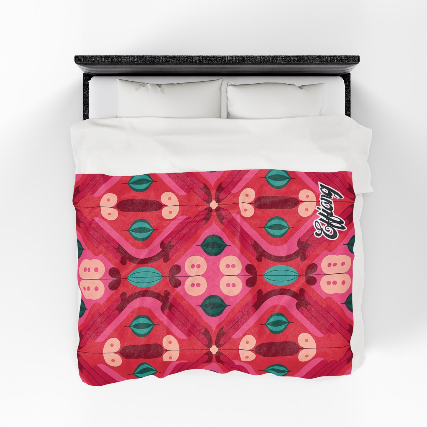 A bed with a white pillow and a vibrant red duvet featuring a symmetrical, African-inspired blanket design in pink, red, teal, and green. The word Lovely is written in white script in the top right corner of the duvet. Viewed from above.