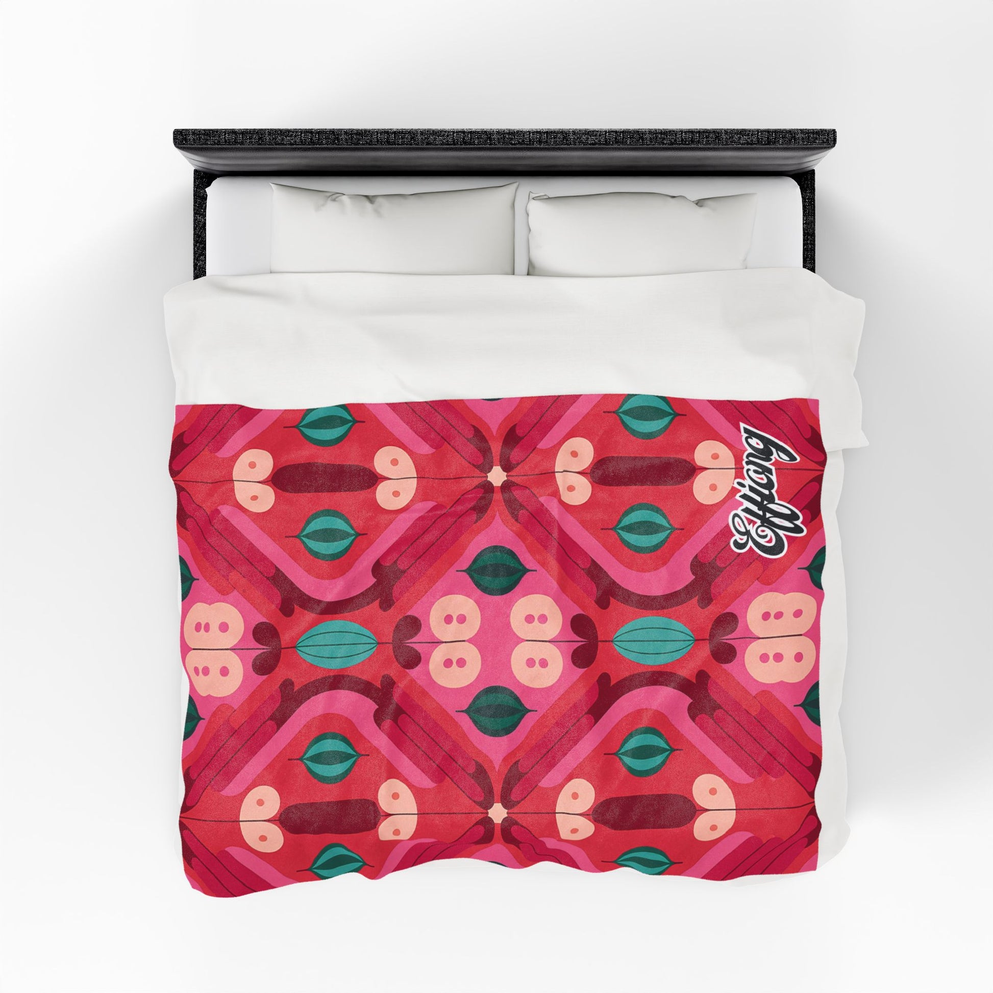 A bed with a white pillow and a vibrant red duvet featuring a symmetrical, African-inspired blanket design in pink, red, teal, and green. The word Lovely is written in white script in the top right corner of the duvet. Viewed from above.