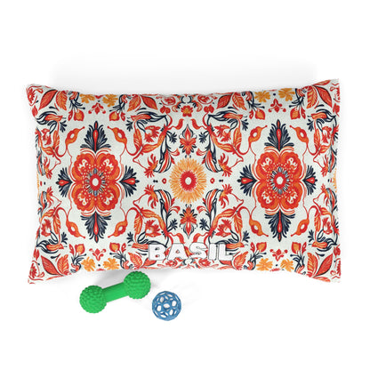 A rectangular pillow with “BASIL” in white block letters and an African-inspired bold floral pattern in red, orange, black, and cream. In front of the pillow are two dog toys: a green dumbbell-shaped toy and a small blue ball.
