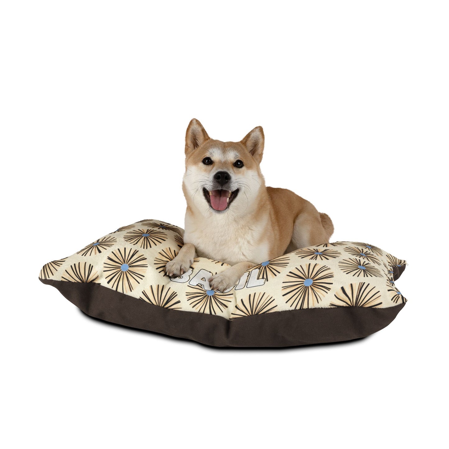 A Shiba Inu dog lies on an African-inspired beige and brown pillow bed with a happy expression, mouth open and tongue out. The bed features a starburst design on top and a solid brown base, set against a plain white background.