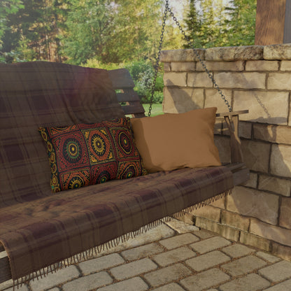 A wooden porch swing with a plaid blanket and weatherproof outdoor pillows—one solid tan, one with a colorful geometric pattern—sits on a stone patio. The customizable design hangs by metal chains, framed by green trees and sunlight in the background.