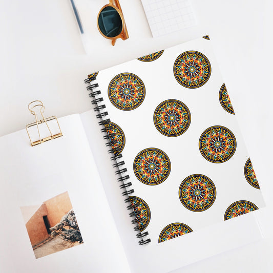 A premium African-inspired spiral notebook with colorful mandala designs on a white cover rests on an open book. Nearby are a gold binder clip, sunglasses, and a grid-patterned notepad. The open book displays a rustic building and rocky ground.