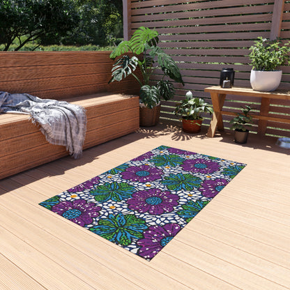 A cozy outdoor deck with wood flooring features a colorful weather-resistant rug with purple and green floral patterns. Surrounding the rug are potted plants, a wooden bench with gray cushions and a plaid throw, a lantern, and horizontal wooden slats.